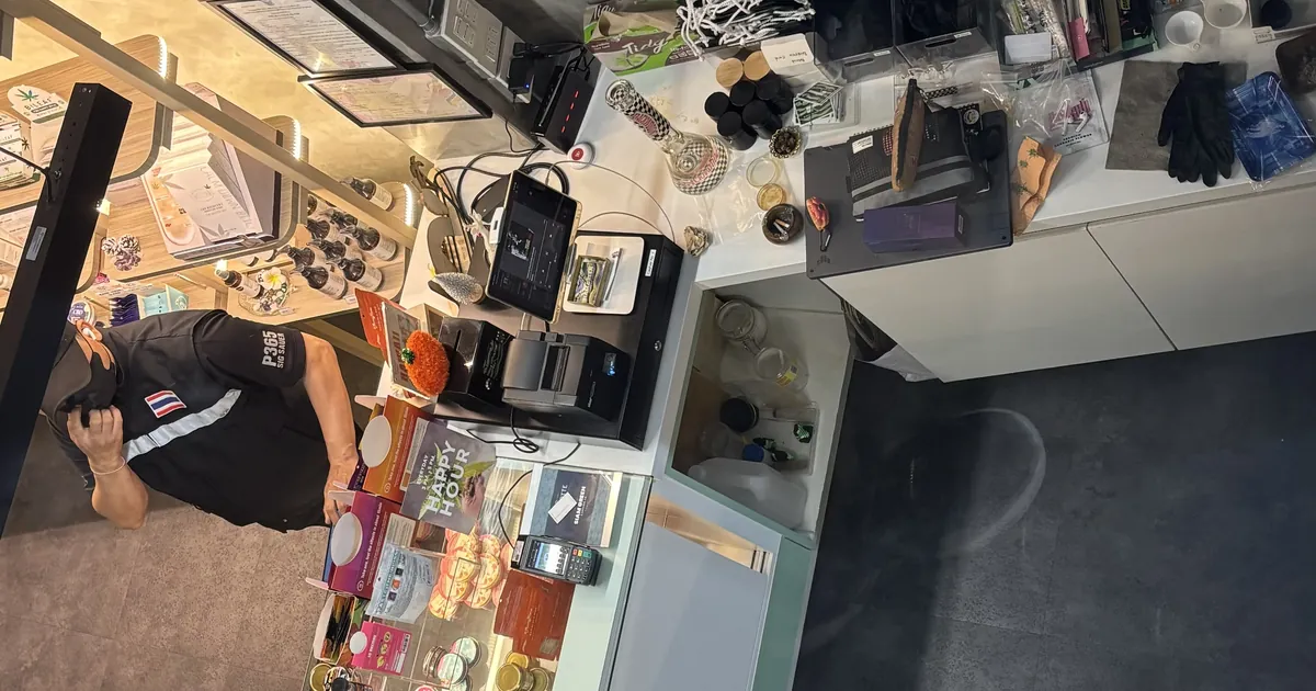 Overhead view of a Bangkok dispensary counter, a staff member at the till among shelves and products