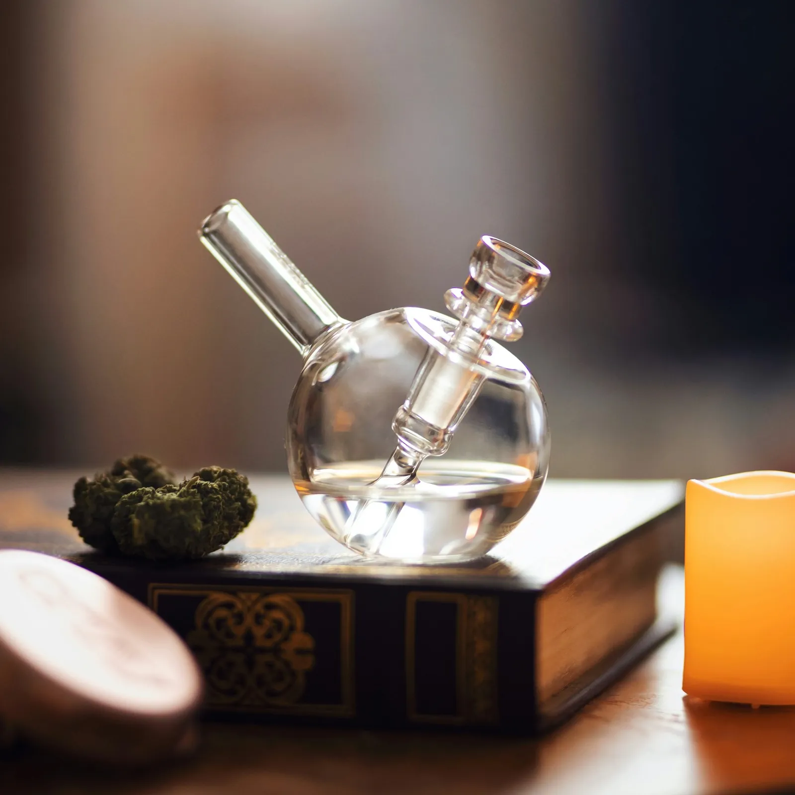 Spherical glass bong resting on an old book beside a glowing candle and cannabis bud - contemplative and atmospheric