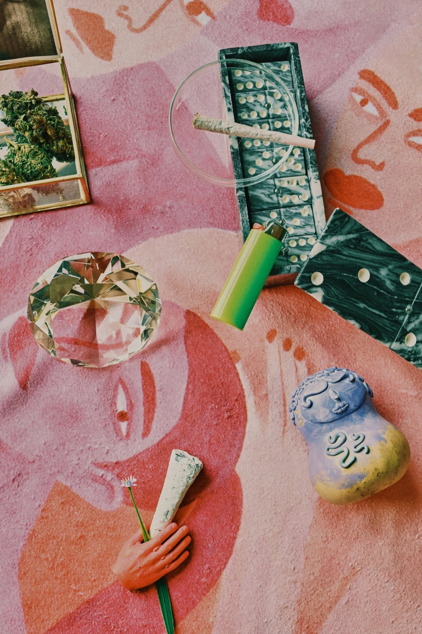 Artistic overhead flat-lay on a painted pink surface: joint, crystal, small Buddha figurine, lighter, framed bud photo - a ritual of wonder