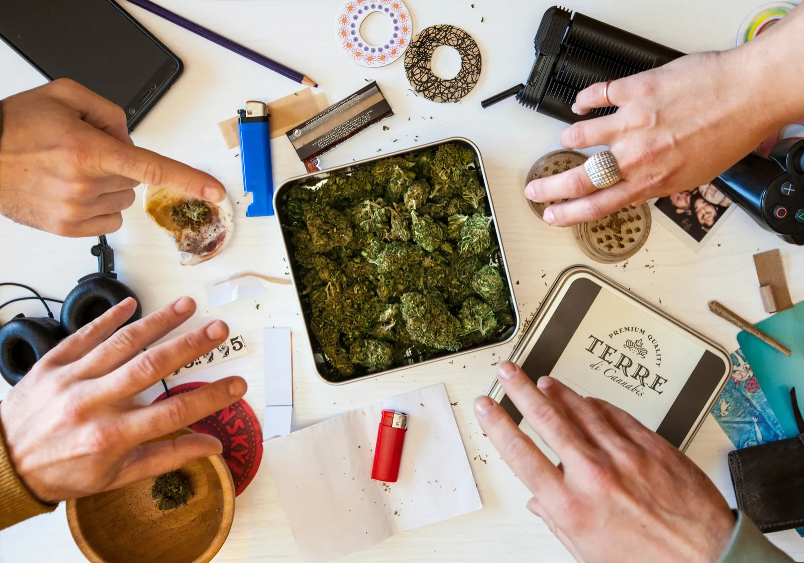 Top-down communal cannabis session with tin, jars, grinder and three hands - Dispensary Ratings Lie Bangkok