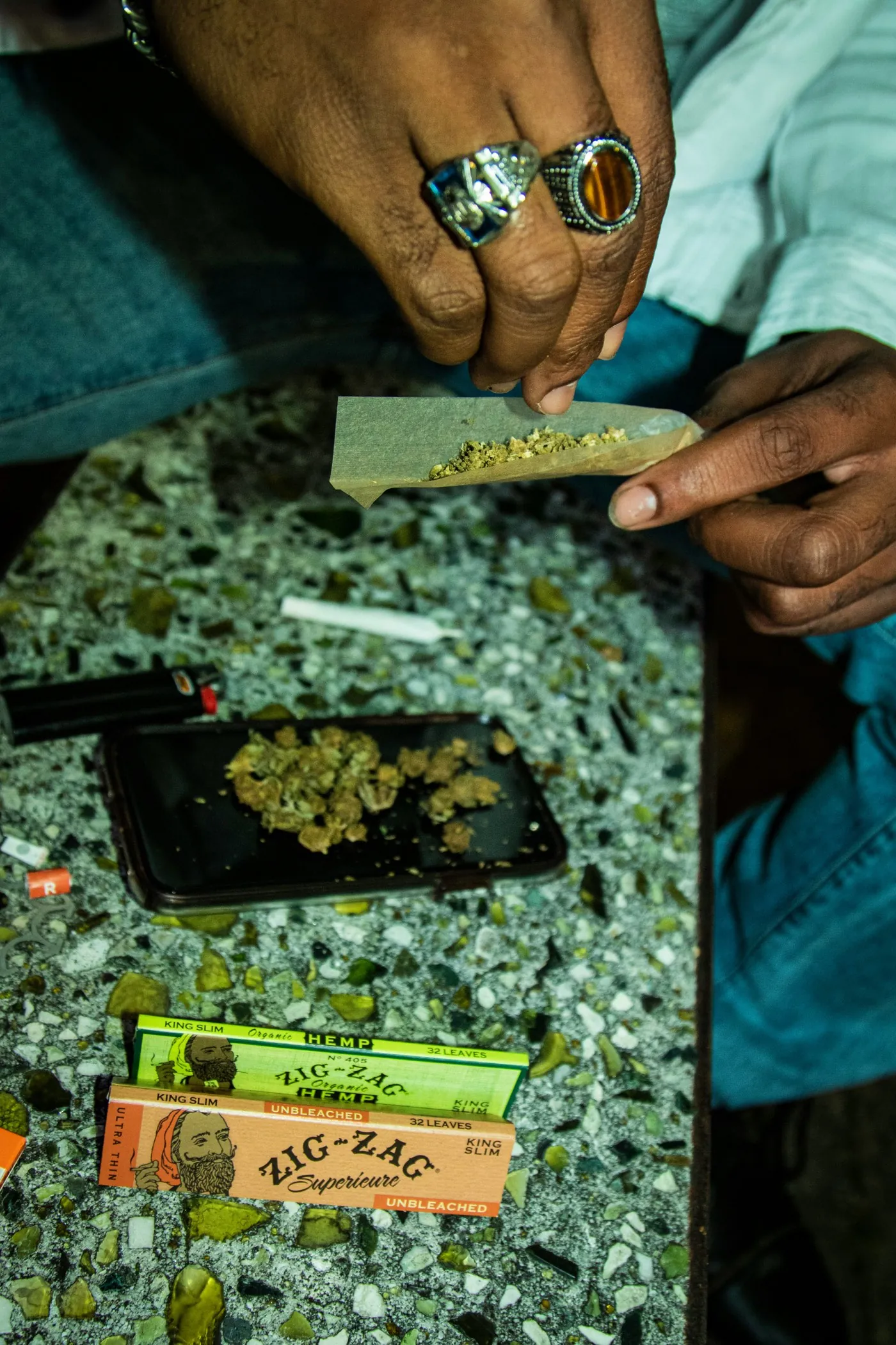 Hands with rings rolling a joint using Zig-Zag papers over a tray of buds