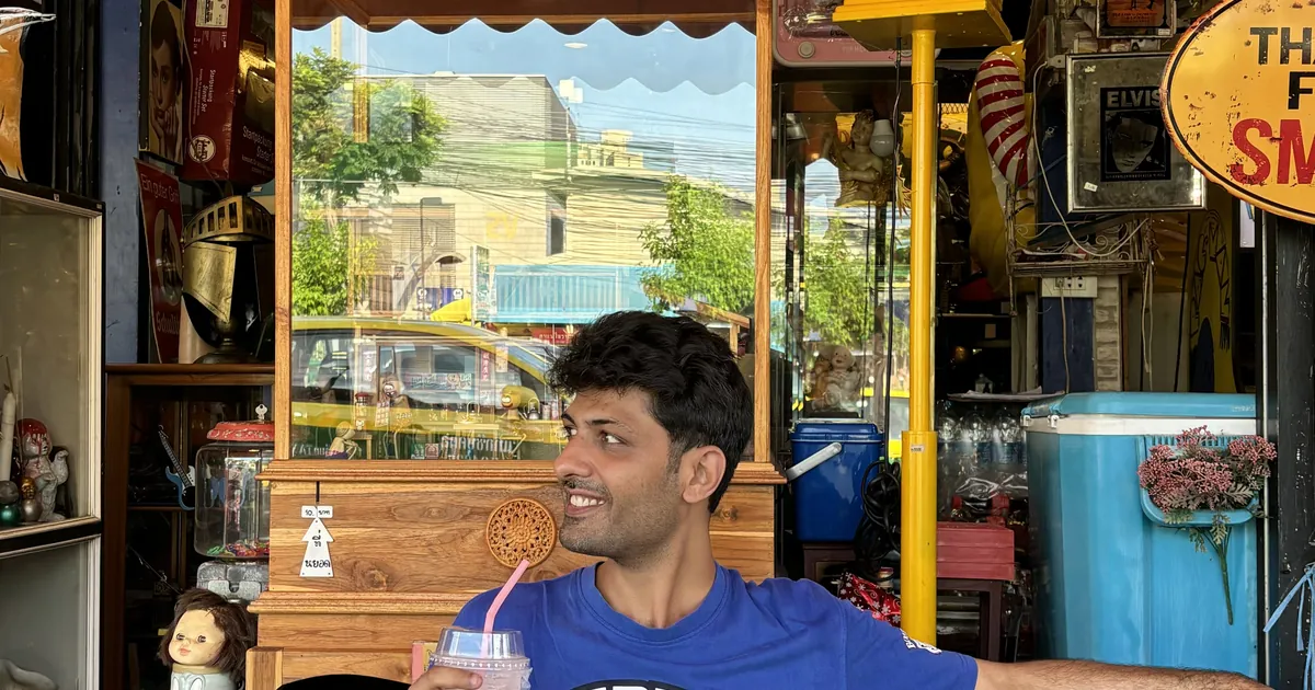 Smiling man with a drink sitting in a colourful shop full of vintage signs, a street behind him